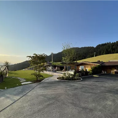 A calm landscape with wooden buildings and a wide meadow. In the background, there are gentle hills and a clear sky.