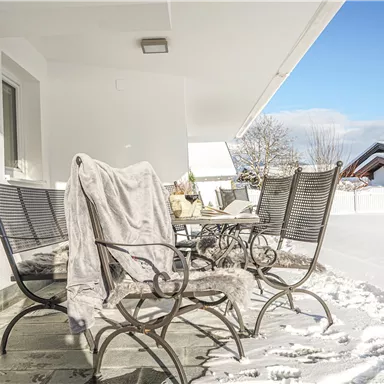 Ein winterlicher Balkon mit weißen Schneedecken. Stühle mit einer Decke und strahlend blauem Himmel im Hintergrund.