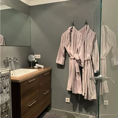 A modern bathroom with a wooden dresser and two fluffy bathrobes. The room features large tiles and a glass shower wall.