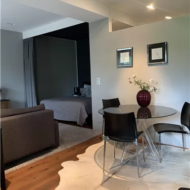 A modern living room with a glass dining table and black chairs. In the background, there is a bedroom and a beautifully decorated rug.