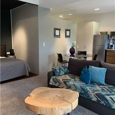 A modern living room with a gray sofa and a round wooden table. In the background, there is a bedroom with a comfortable bed.
