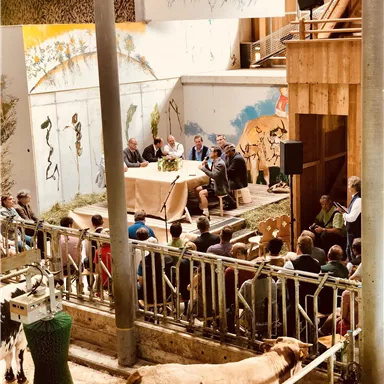 Eine Diskussion findet in einem Agrarzentrum statt. Zuschauer sitzen vor einer Bühne, umgeben von landwirtschaftlichen Elementen und Kühen.