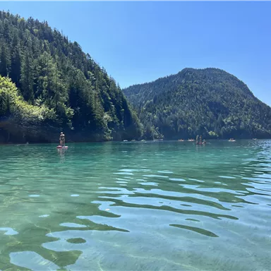 A clear lake surrounded by green forests and mountains. People enjoy the tranquility of the water on a sunny day.