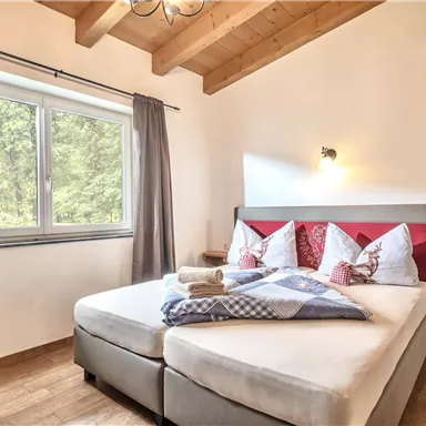 Ein gemütliches Schlafzimmer mit einem Doppelbett und schönen Kissen. Große Fenster lassen viel Tageslicht herein, und die Holzbalkendecke verleiht dem Raum eine warme Atmosphäre.