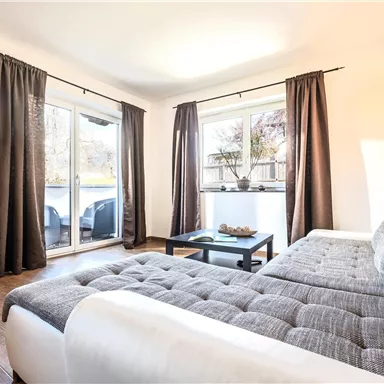 Ein helles Wohnzimmer mit einem gemütlichen Sofa und einem Tisch. Große Fenster mit Vorhängen bieten einen Blick auf den Außenbereich.