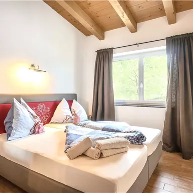 Ein gemütliches Schlafzimmer mit einem Doppelbett und hellen Holzdetails. Große Fenster mit Vorhängen und dekorativen Kissen auf dem Bett sorgen für eine einladende Atmosphäre.