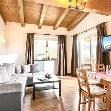Ein helles und modernes Wohnzimmer mit Holzbalkendecke. Es gibt ein gemütliches Sofa, einen Essbereich und große Fenster mit Aussicht.