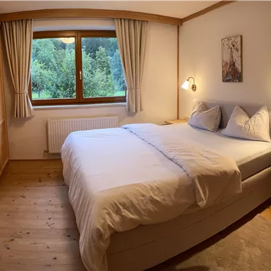 Ein gemütliches Schlafzimmer mit einem großen Boxspringbett, sanften Farben und Holzdetails. Durch das Fenster ist ein Blick ins Grüne sichtbar.