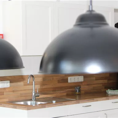 A modern kitchen with wood details and elegant black pendant lights. In the background, dishes and appliances are visible.