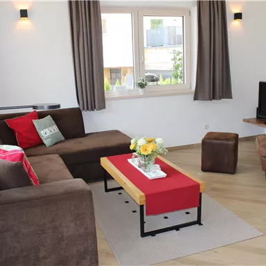 A cozy living room with a brown sofa and a red table. There is a television and plenty of daylight coming through the windows.