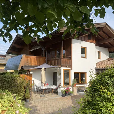Ein gemütliches Haus mit einem großen Balkon beziehungsweise Terrasse  und einem Garten. Im Hintergrund sind Bäume und die Berge sichtbar.
