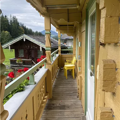 A wooden balcony with yellow chairs and colorful flowers. In the background, wooden houses and green meadows can be seen.