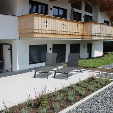 A modern house with a balcony and a terrace. On the terrace, there are reclining chairs, and they are surrounded by a beautiful garden.