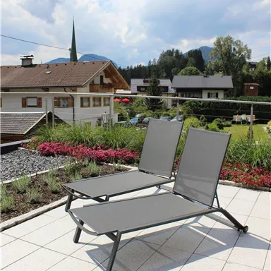 Two gray loungers on a terrace with blue sky and clouds. In the background, houses and trees are visible.