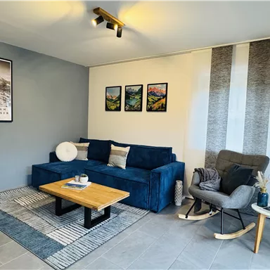 A modern living room with a blue sofa and a wooden coffee table. The walls are decorated with pictures and there is a cozy armchair.