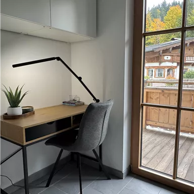 A modern study with a wooden desk and an ergonomic chair. A beautiful outdoor view with trees and a terrace can be seen through the window.