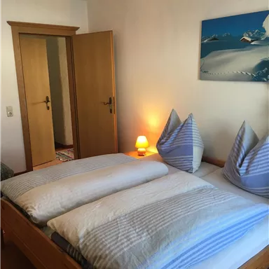 A cozy bedroom with a double bed and blue pillows. There is a warm glow of light and a picture of mountains on the wall.