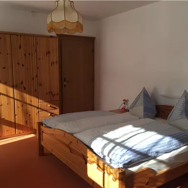 A simple bedroom with a large bed and two pillows. In the background, there is a wooden wardrobe and the room is brightly lit.