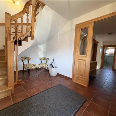 An inviting hallway with a wooden staircase and two chairs. The room is brightly lit and has brown tiles.