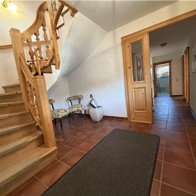 An inviting hallway with a wooden staircase and tiled floor. On the left side, there are some chairs and a door that leads to additional rooms.