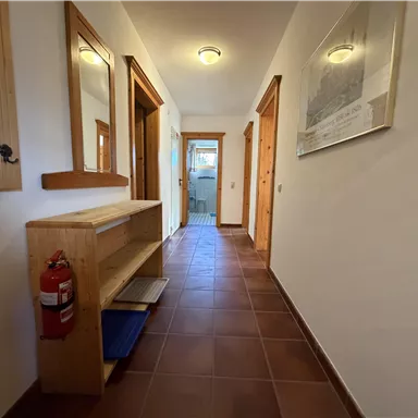 A bright hallway with wooden paneling and mirrors. On the left side, there is a shoe rack and a fire extinguisher is hanging on the wall.