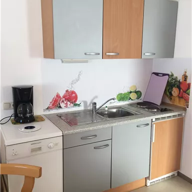 A small kitchenette with gray and wooden fronts. There is a sink, a stove, and a coffee machine, as well as a wall design featuring vegetables and fruit.