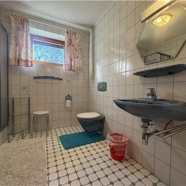 A bathroom with tiled walls and a large window. There is a shower, a sink, and a toilet, as well as some bathroom accessories.
