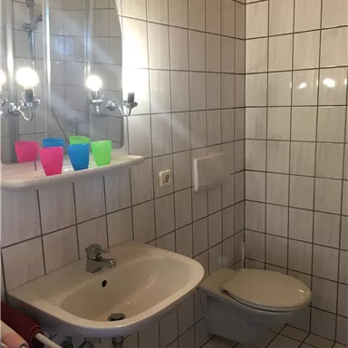 A simple bathroom with white tiles and a sink. On the shelf, there are colorful cups, and there is mirror lighting.