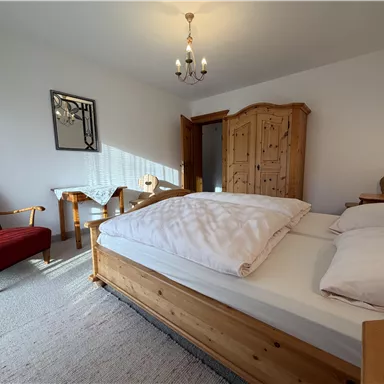 A cozy bedroom with a large bed and a wooden wardrobe. A red armchair and a small table complete the decor.