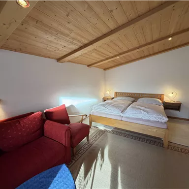 A cozy bedroom with a double bed and a red sofa. The wooden ceiling and soft lighting create an inviting atmosphere.