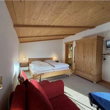 A bright bedroom with wooden ceilings and a large bed. There is a red couch and a television on the wall.