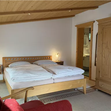 A cozy bedroom with a large bed, two nightstands, and a red sofa. The walls are bright, and there is a rustic wooden wardrobe.