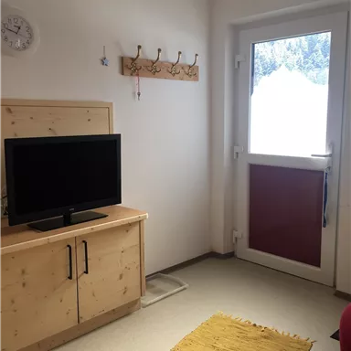 A cozy room with a television and a wooden cupboard. A rug lies on the floor and a door leads outside.