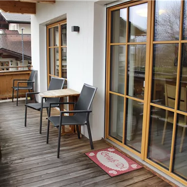 A cozy balcony with wooden floorboards, two chairs, and a table. The large windows offer a beautiful view outside.