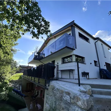 Ein modernes Haus mit Balkon, umgeben von grünen Bäumen. Der Himmel ist klar und blau, was eine ruhige Umgebung schafft.