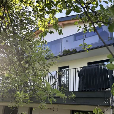 Ein modernes Haus mit einem Balkon und großen Fenstern. Die Umgebung ist von grünen Bäumen umgeben.