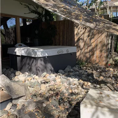 A hot tub sits on a bed of stones, surrounded by plants and wood. In the background, sunny trees and a brightly lit terrace can be seen.