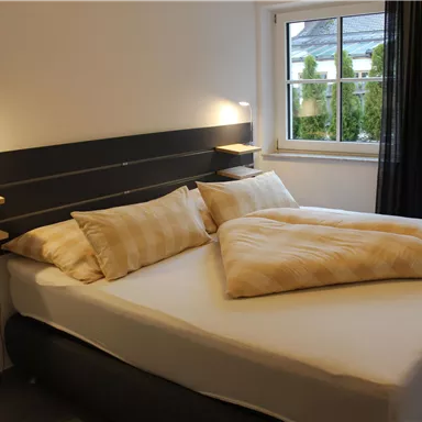 A modern bedroom with a large double bed and two pillows. Large windows provide plenty of daylight and a view of the outdoors.