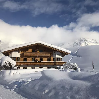 A cozy wooden house in the snow, surrounded by a wintry landscape. The mountains are visible in the background, and the sky is partly cloudy.
