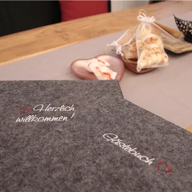 Ein schön gedeckter Tisch mit einem grauen Menübuch und einer herzlichen Willkommensbotschaft. Im Hintergrund sind gefüllte Tüten und dekorative Elemente angeordnet.