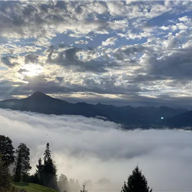 A breathtaking view of mountains shrouded in fog. The sky is filled with clouds and rays of sunshine.