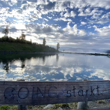 A wooden bench by the shore of a calm body of water with the inscription "GOING strengthens". In the background, clouds are reflected in the water, and the landscape is softly illuminated.