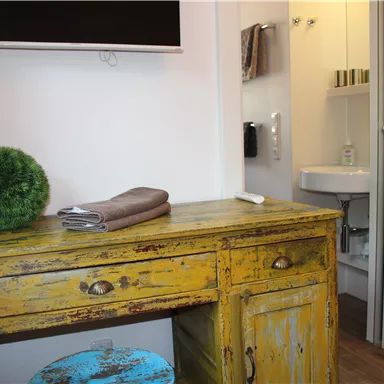 A cozy room with a yellow vintage piece of furniture and a television on the wall. In the background, a small bathroom with a sink can be seen.