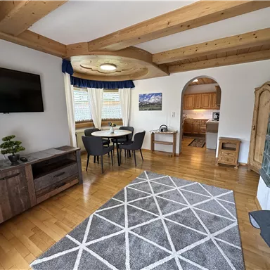 Ein gemütlicher Wohnraum mit Holzbalkendecke und einem großen Fenster. Der Raum ist mit einem Esstisch, einem Sofa und einem Teppich ausgestattet.