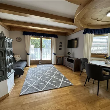 Ein gemütliches Wohnzimmer mit Holzbalkendecke und großen Fenstern. Es gibt eine Couch, einen Tisch und einen modernen Fernseher.