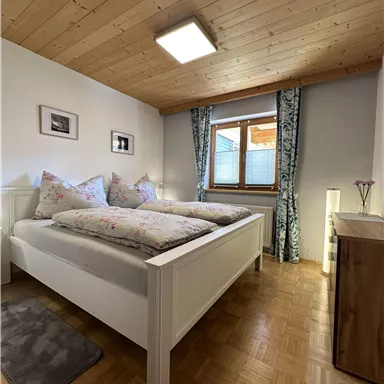 Ein gemütliches Schlafzimmer mit einem Doppelbett und Holzdecke. Der Raum hat ein Fenster mit Vorhängen und ist hell und einladend.