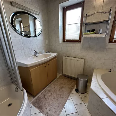 Ein modernes Badezimmer mit einer Duschkabine, einer Badewanne und einem Doppelwaschbecken. Helle Fliesen und Fenster sorgen für eine freundliche Atmosphäre.