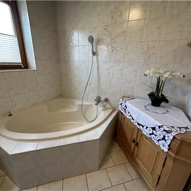 Ein gemütliches Badezimmer mit einer modernen, eckigen Badewanne und einer Dusche. Neben der Badewanne steht eine Holzkommode mit einer dekorativen Pflanze.