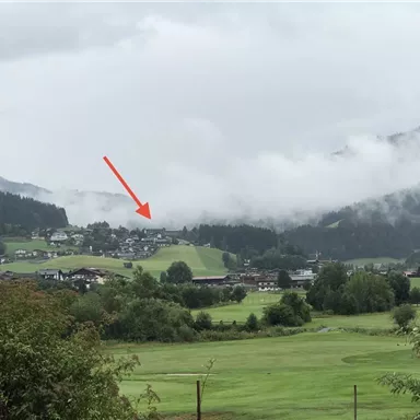 Eine malerische Landschaft mit Hügeln und einem kleinen Dorf im Hintergrund. Der Himmel ist bewölkt, und die Umgebung wirkt ruhig und grün.