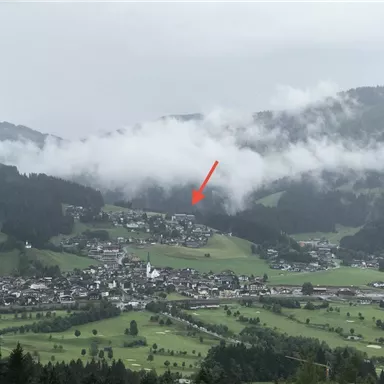 Ein malerisches Dorf in den Bergen, umgeben von grünen Wiesen und niedrig hängenden Wolken. In der Mitte ist eine Kirche sichtbar.